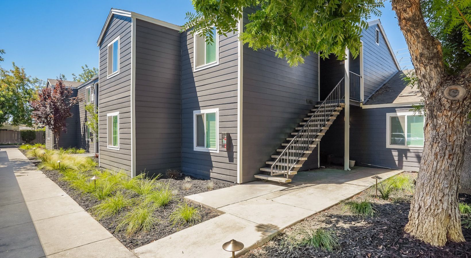 Two-story gray apartment building with outdoor stairs and a tree, on a sunny day.