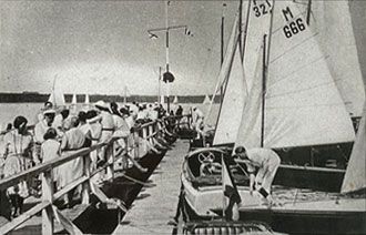 Ein Schwarzweißfoto von Leuten, die auf einem Dock sitzen und Segelboote beobachten