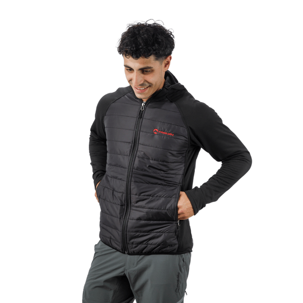 Hombre con chaqueta negra y pantalón gris de pie, con las manos en los bolsillos, mirando hacia abajo y sonriendo.