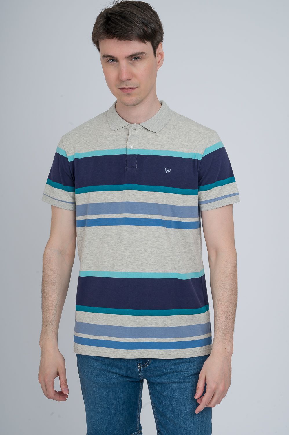 Persona de pie frente a un fondo liso, vestida con una camisa polo a rayas y pantalones vaqueros.