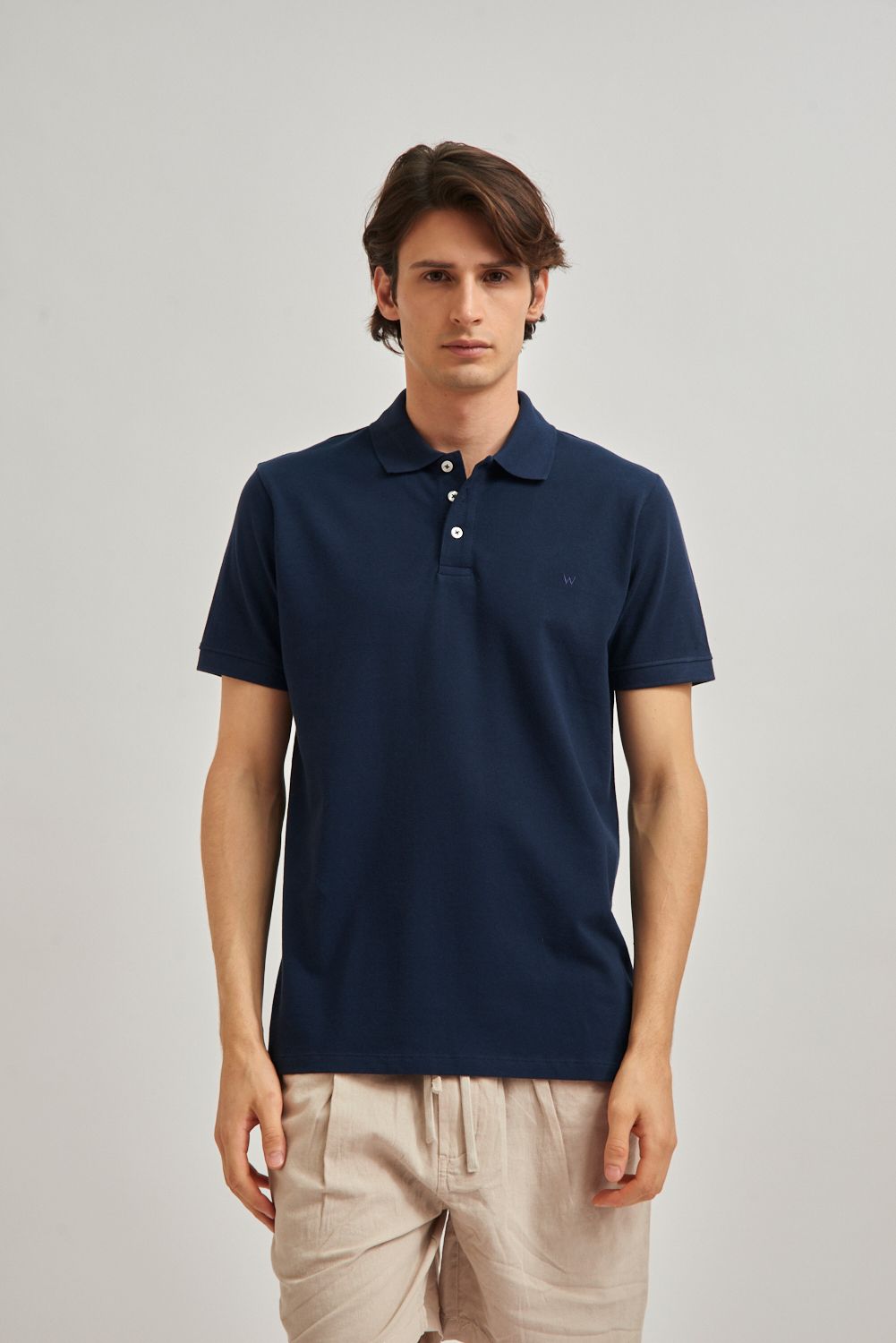 Hombre con polo azul claro y pantalones cortos beige, de pie sobre un fondo gris liso.