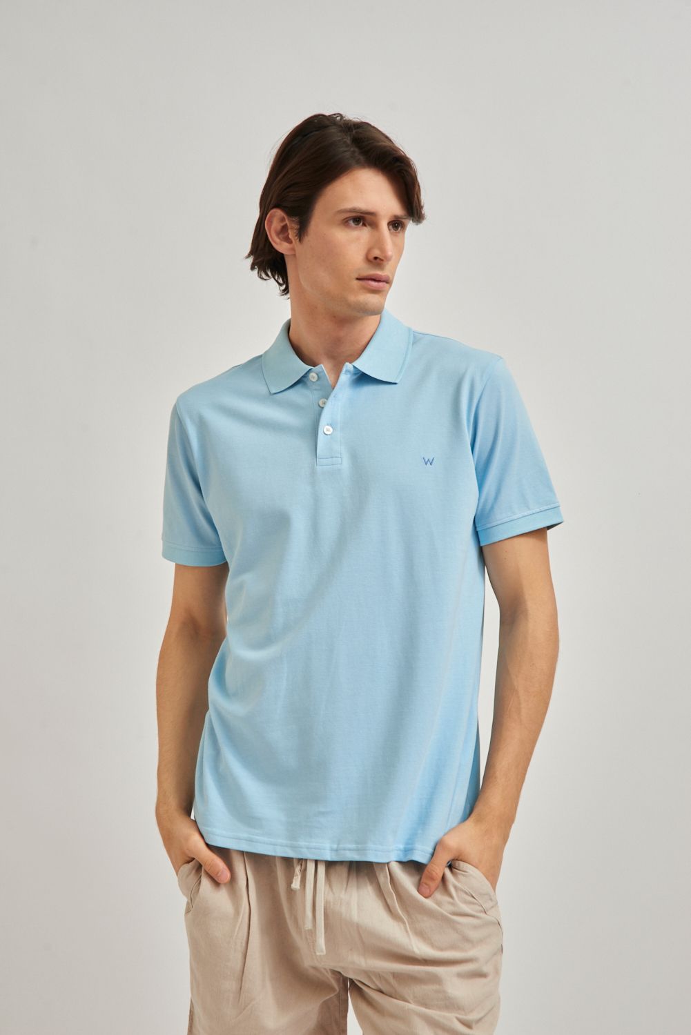 Hombre con polo azul claro y pantalones cortos beige, de pie sobre un fondo gris liso.