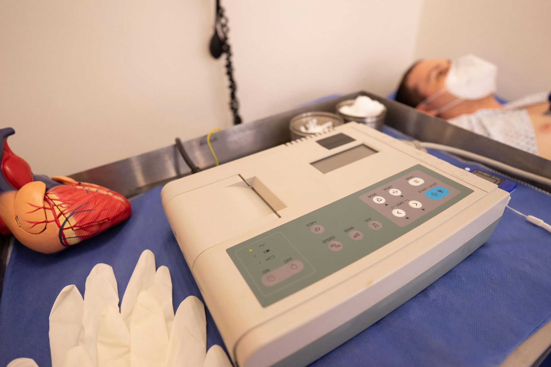 Medical exam table with a blood pressure monitor, gloved hand, and a patient lying in the background