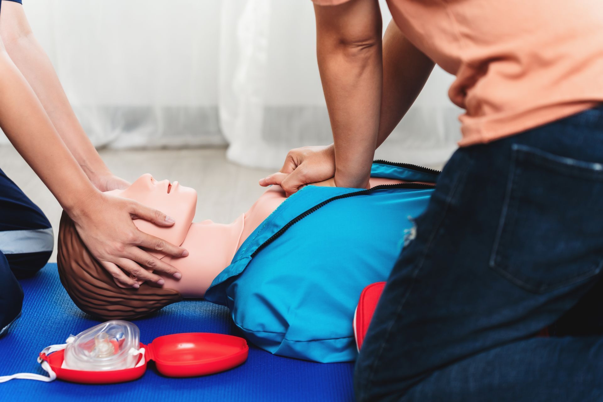 Two people performing CPR on a person lying on the floor beside a red AED kit.