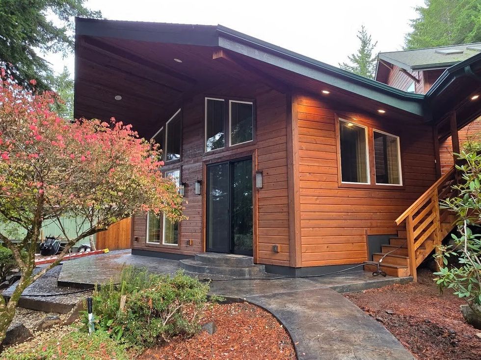 Modern wooden cabin with large windows, black stone path, and red shrubs in a wooded setting