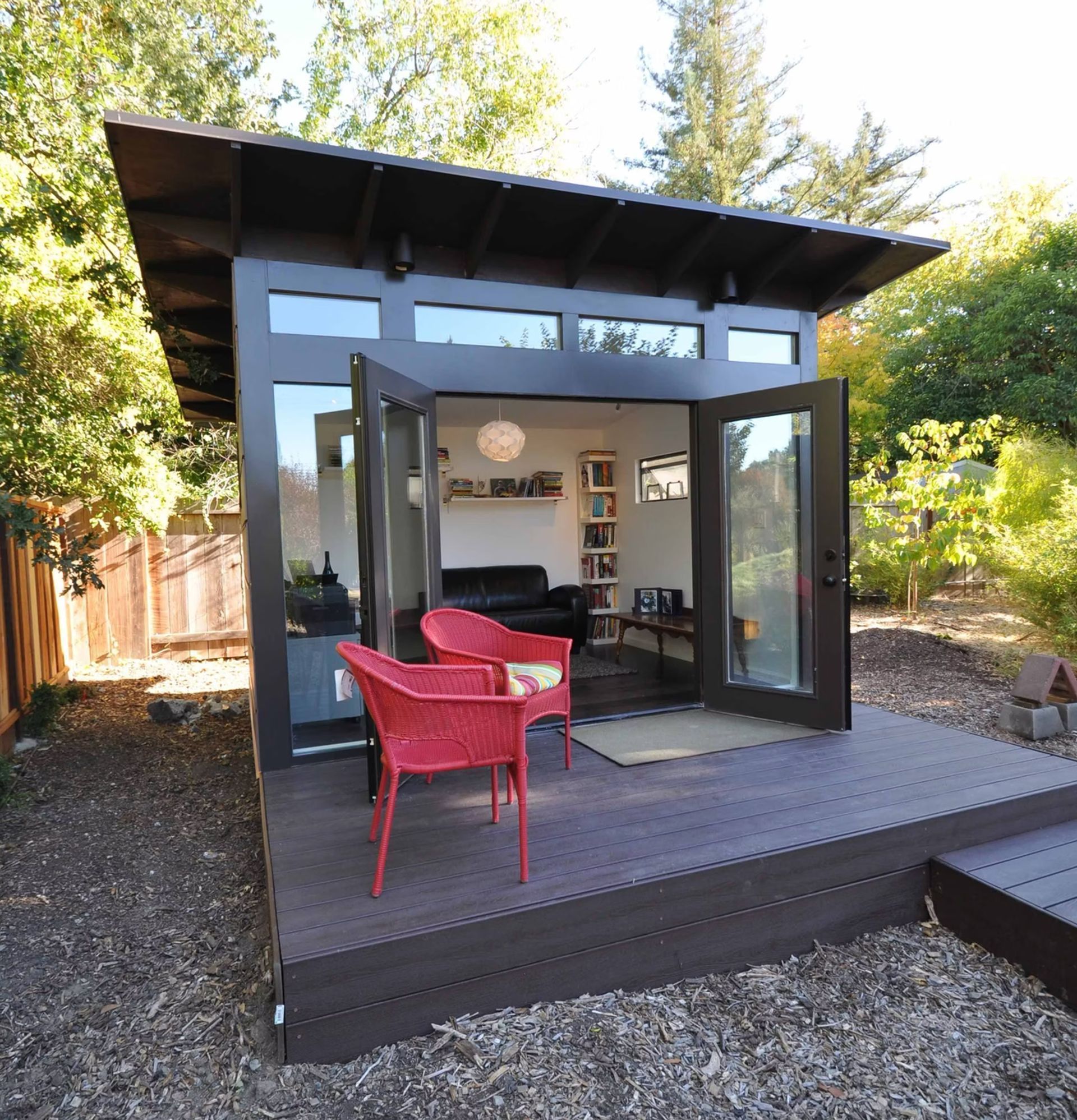 Modern tiny house with open glass doors, red chair on a wooden deck, surrounded by trees