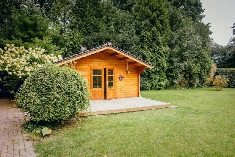 Small orange wooden garden shed with double doors in a grassy yard, surrounded by trees and shrubs