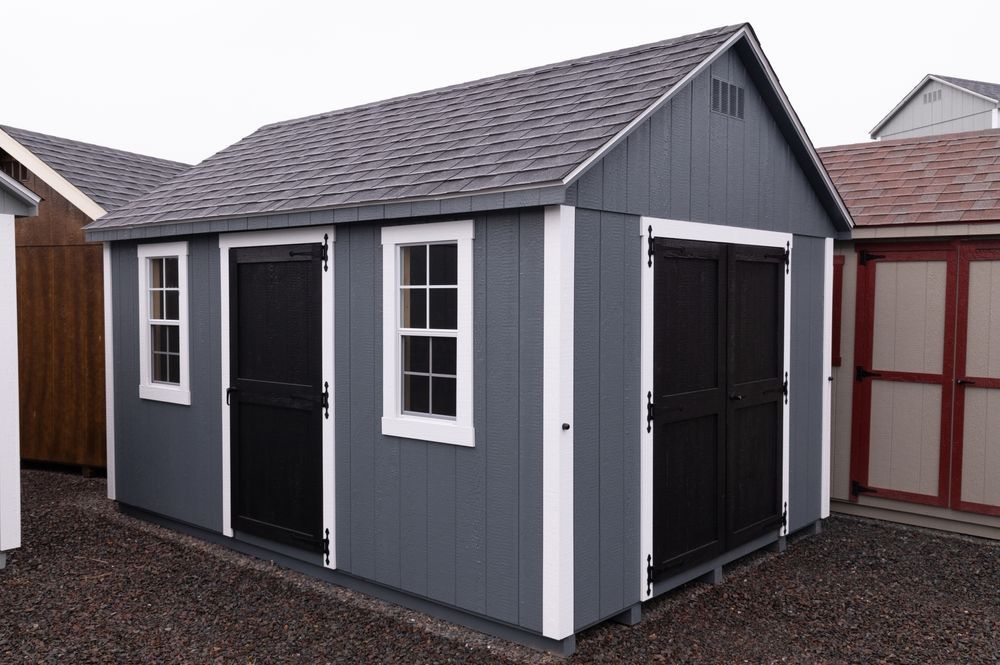Small gray shed with black double doors and white-trimmed windows on a gravel lot