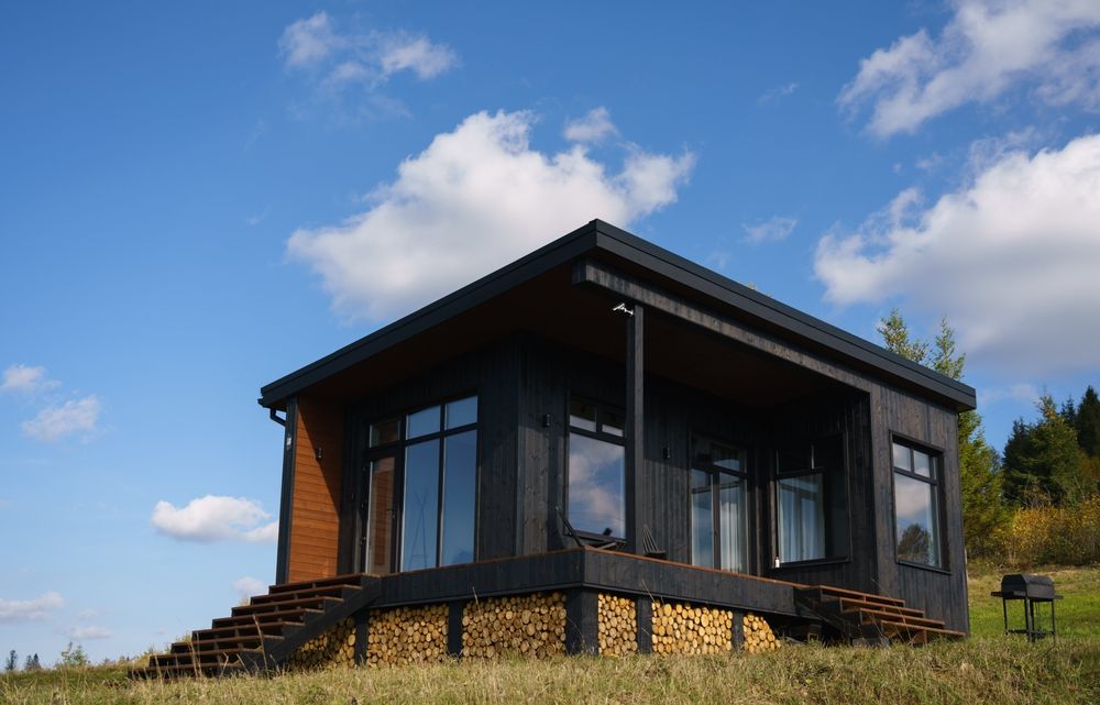 Modern dark wood cabin on a grassy hillside under a blue sky with clouds