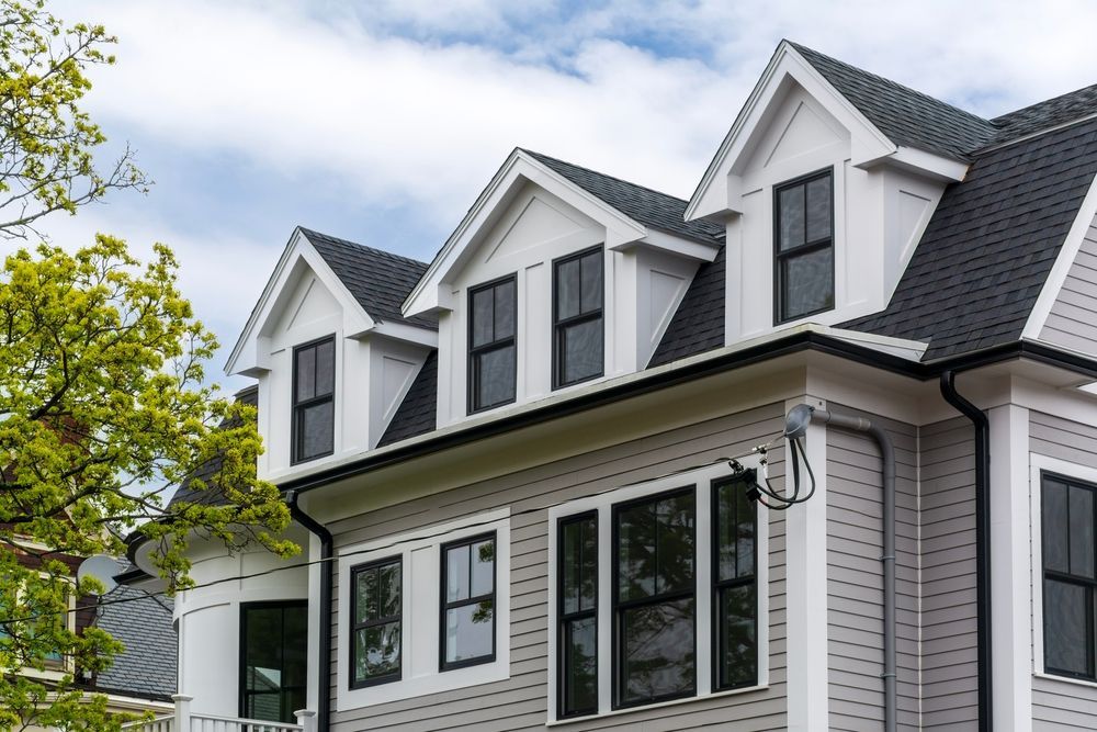 White two-story house with black roof, dormer windows, and large front windows beside a leafy tree
