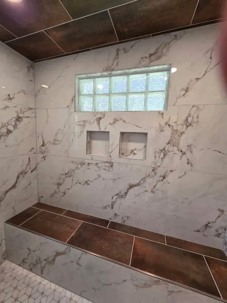 Empty tiled shower with marble-look walls, dark brown bench and ledge, and small frosted glass blocks.