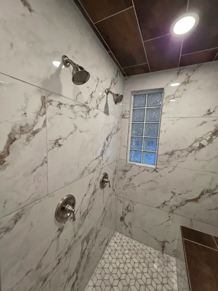 Empty shower stall with marble-look tile, chrome fixtures, and a small frosted window.