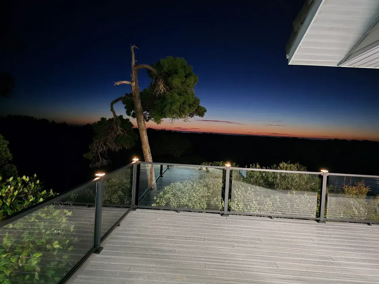 Patio at dusk overlooking water, with glass railing, tree silhouette, and twilight sky.