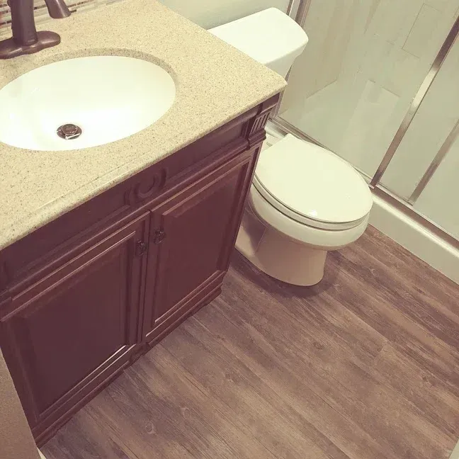 Bathroom with wooden vanity, beige countertop sink, toilet, and glass shower on wood-look floor.