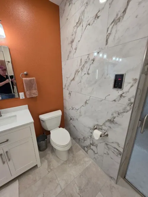 Small bathroom with white toilet, marble tile shower wall, orange accent wall, and vanity sink.