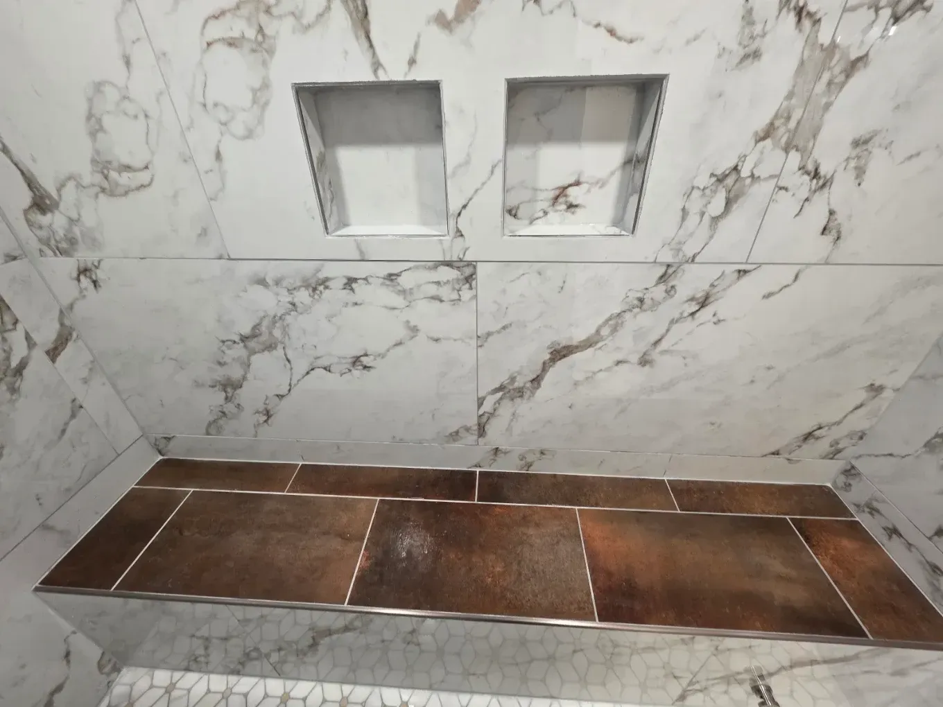 Marble-tiled shower niche above a brown tiled bench in a bathroom.