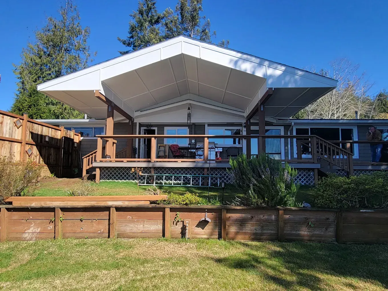 Modern house with a large white roof, wooden deck, and fenced backyard under a blue sky