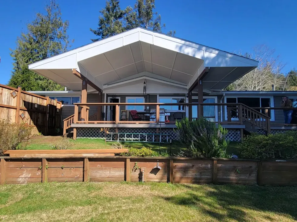 Modern house with a large white roof, wooden deck, and fenced backyard under a blue sky