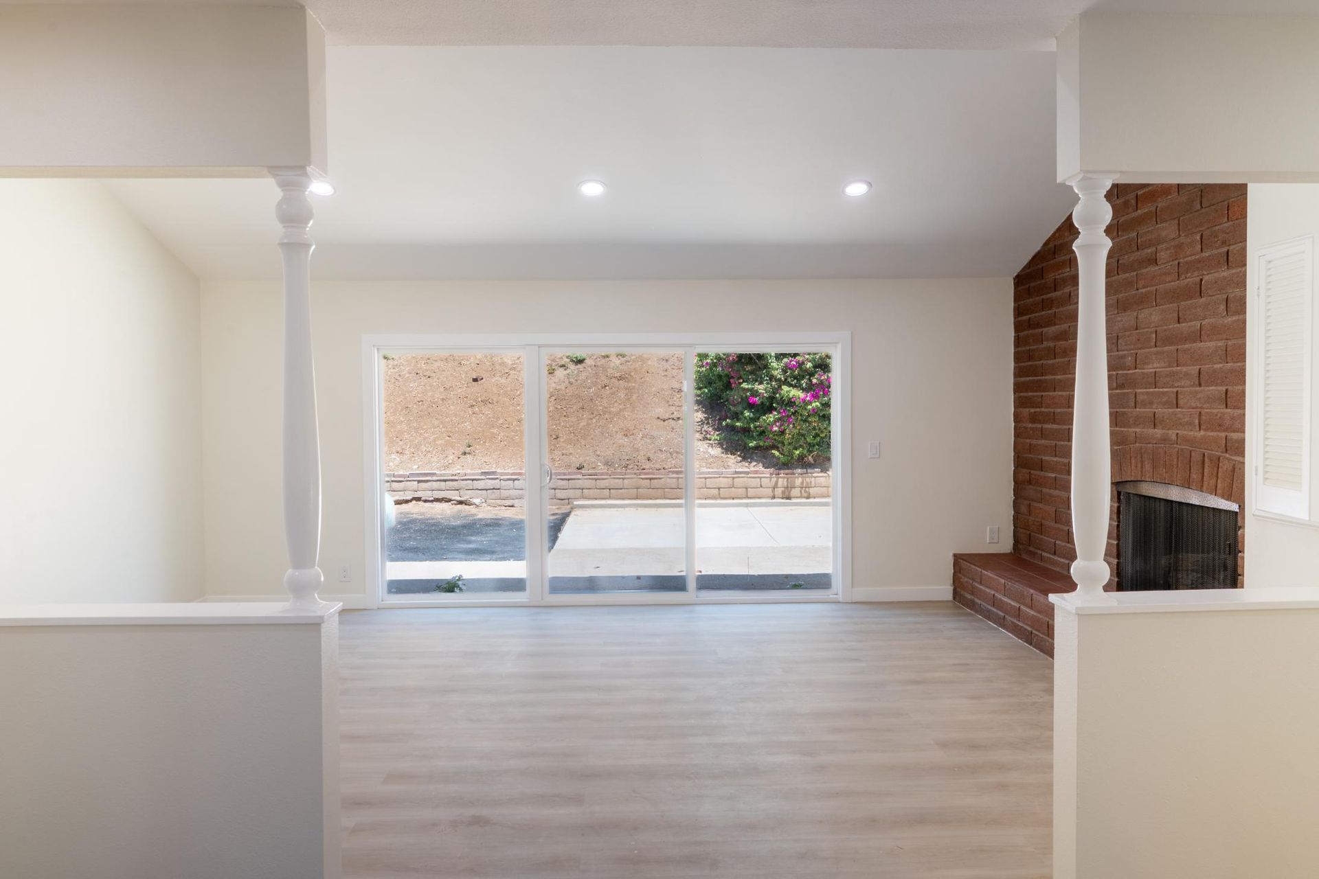 Bright empty living room with brick fireplace, white columns, and large windows overlooking a patio.