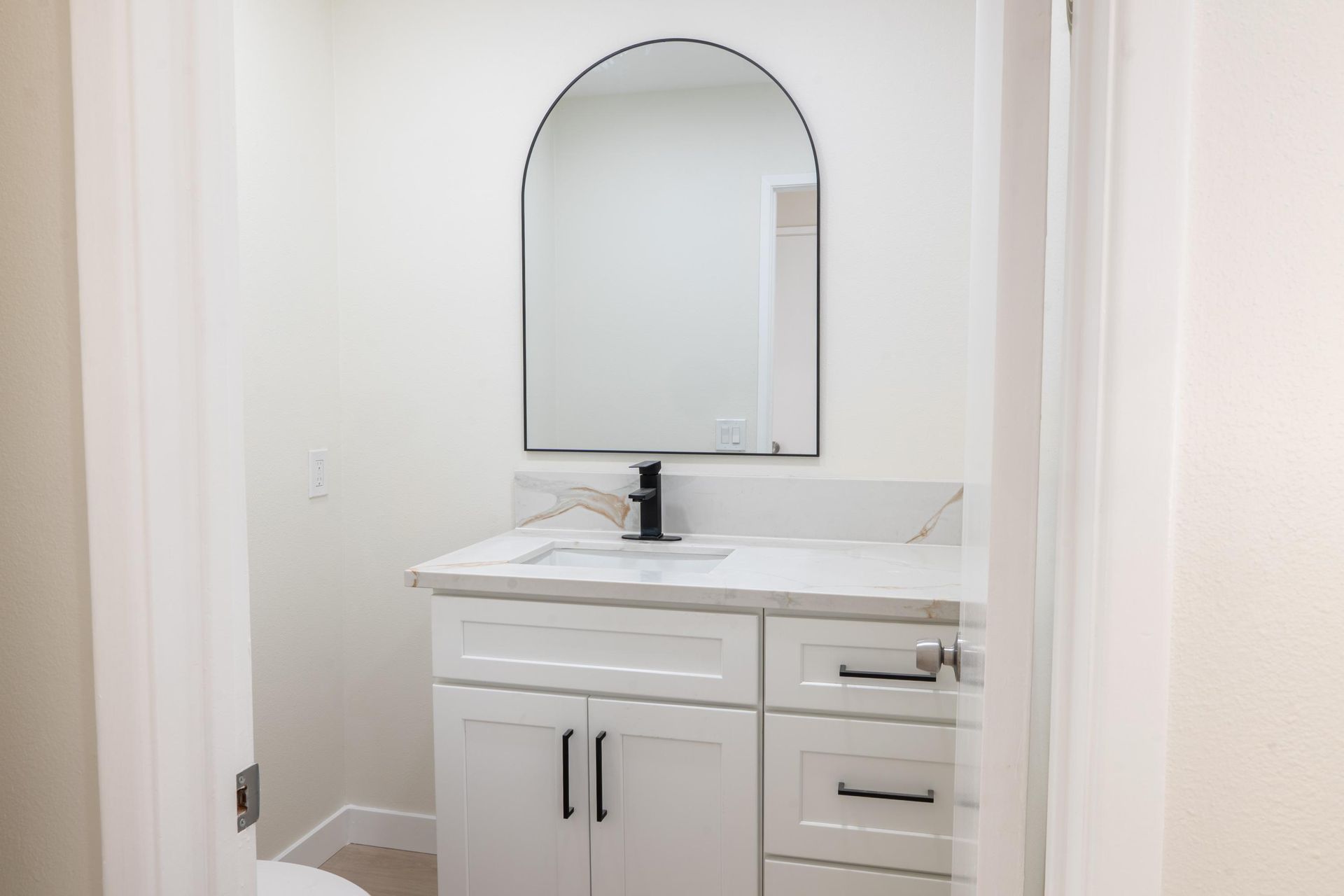 Small white bathroom vanity with arched mirror, black faucet, and light countertops in a narrow alcove
