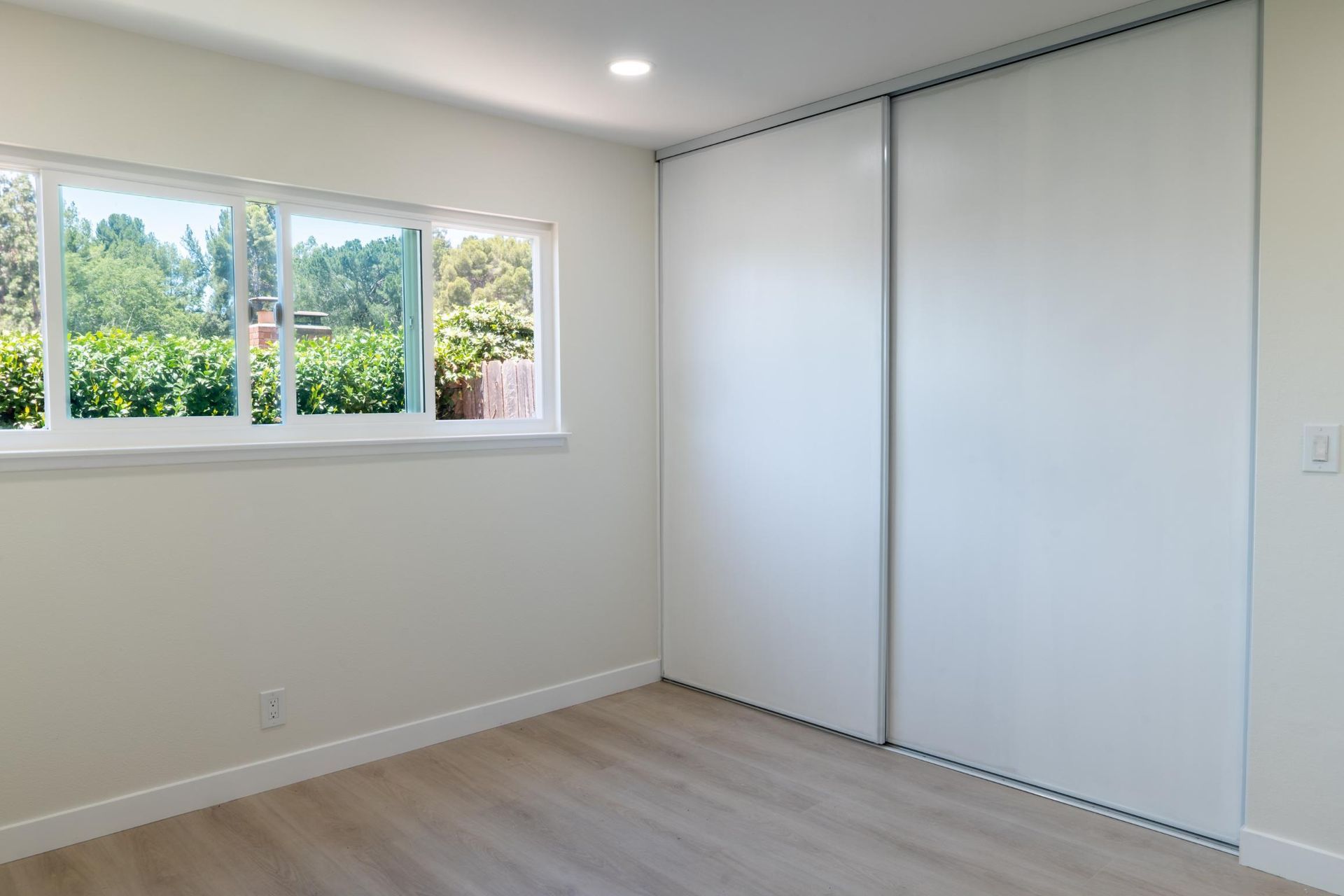 Empty white bedroom with closet doors, wood floor, and a window overlooking trees.