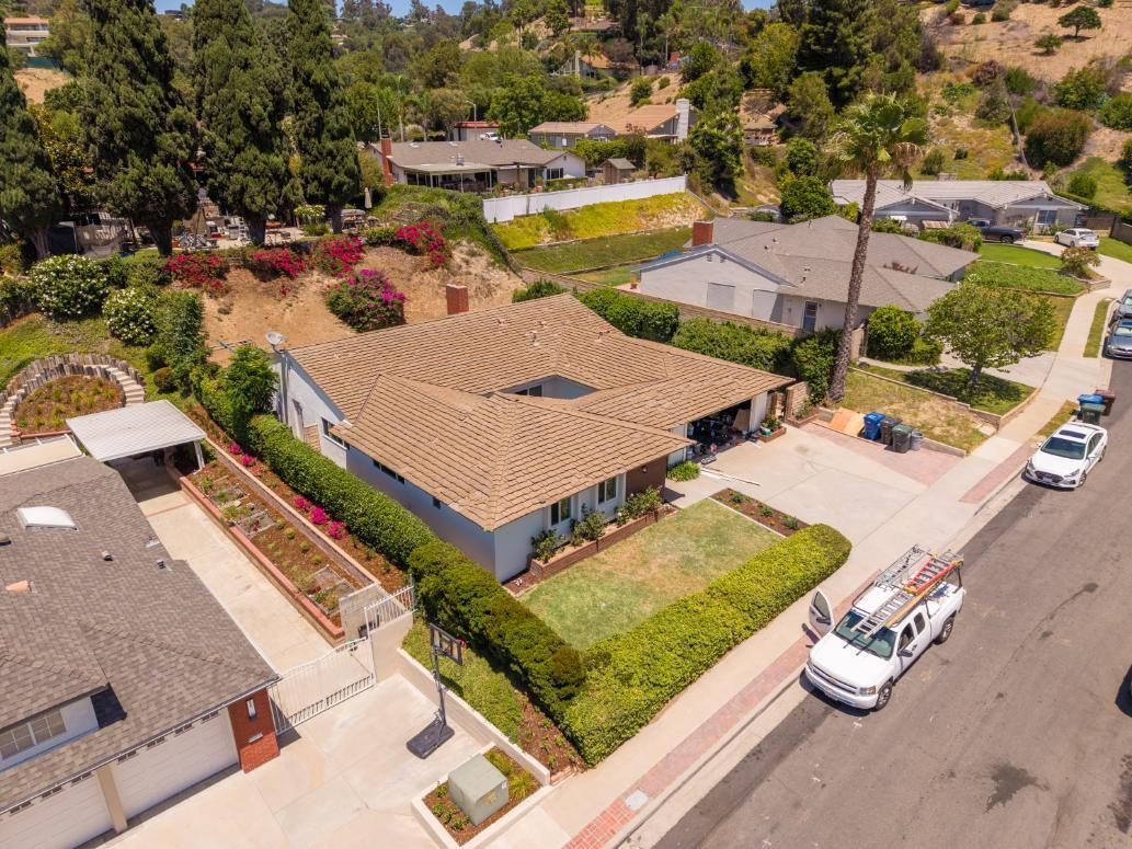 Aerial view of a suburban house with a tan roof, hedges, and parked cars along a quiet street.