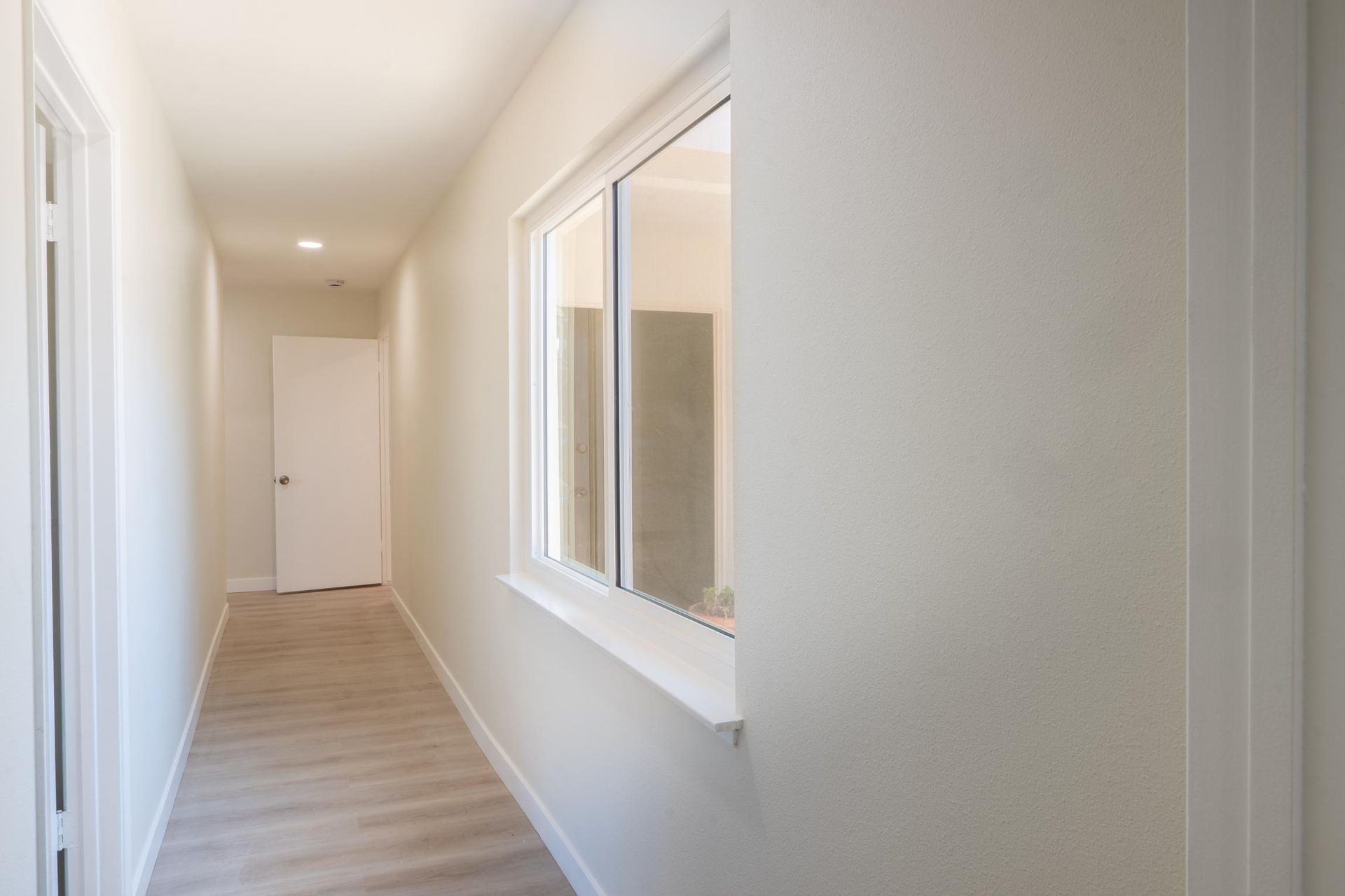 Bright narrow hallway with white walls, wood floor, and a window on the right