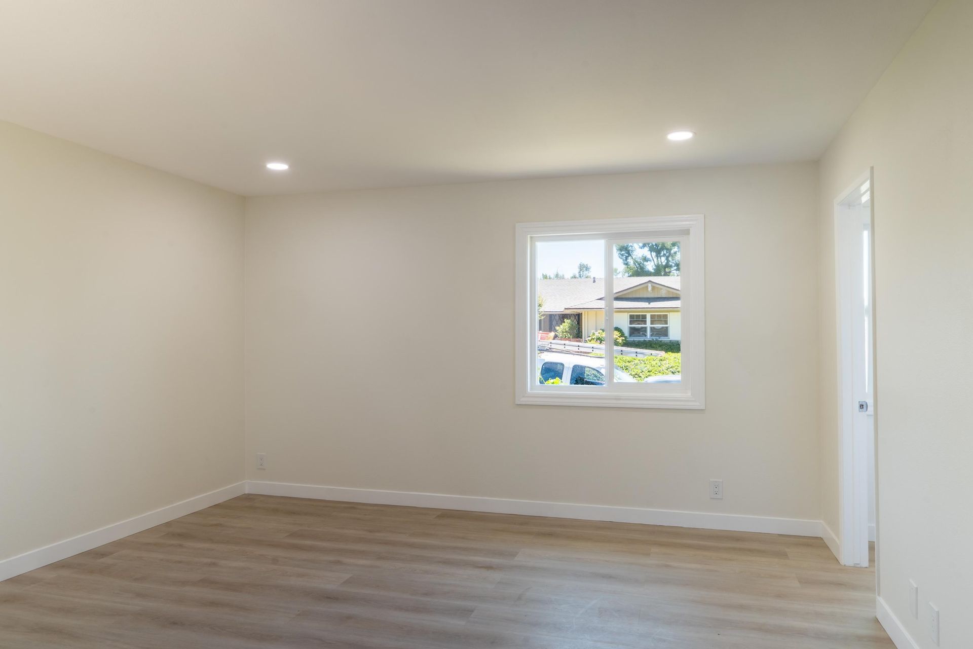 Empty beige room with hardwood floor, recessed light, and a small window overlooking houses.