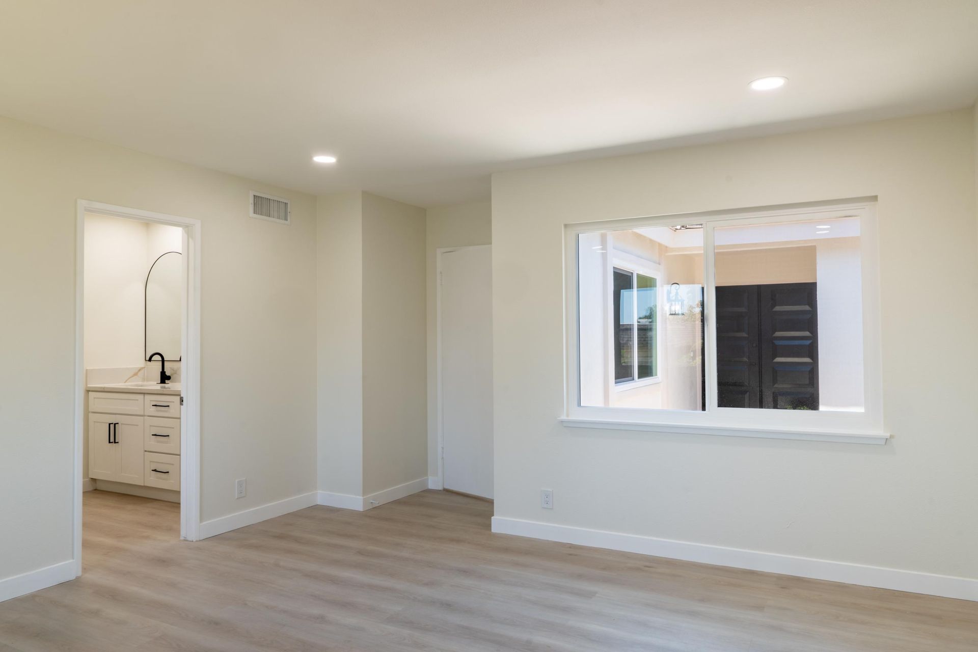 Empty beige bedroom with wood floors, window, and open doorway to bathroom