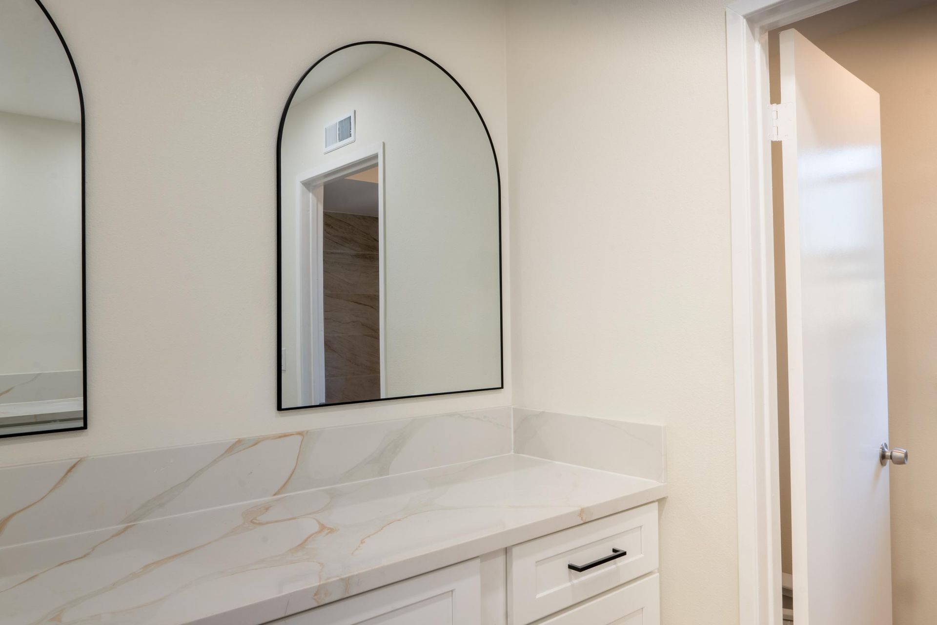 White bathroom vanity with marble countertop and arched mirror beside an open doorway