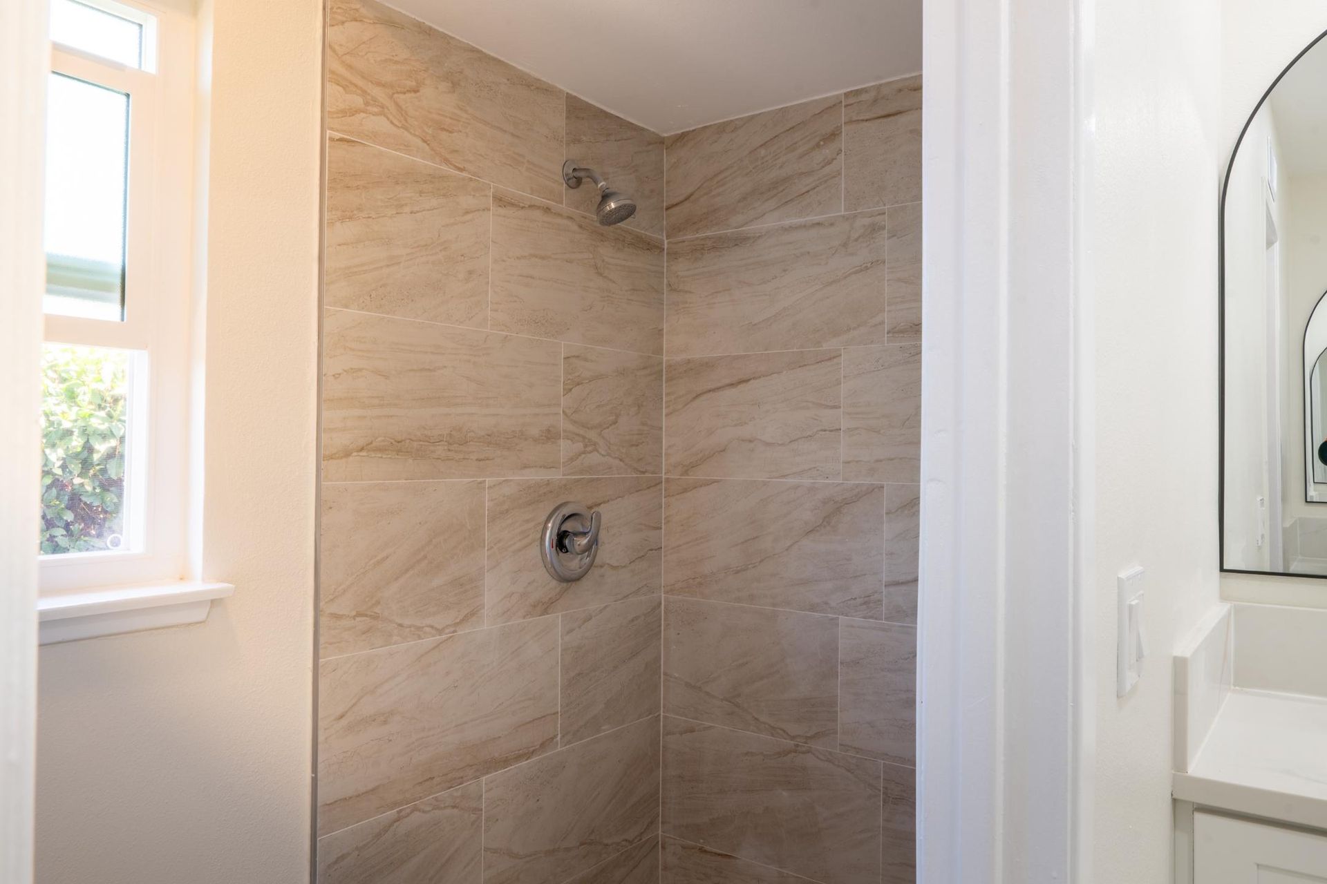 Walk-in shower with beige tile walls and chrome fixtures beside a bathroom vanity and window