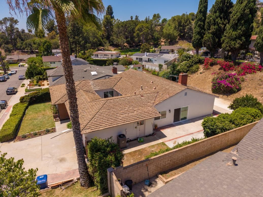 Aerial view of a suburban house with tan roof, driveway, and surrounding trees and neighboring homes.