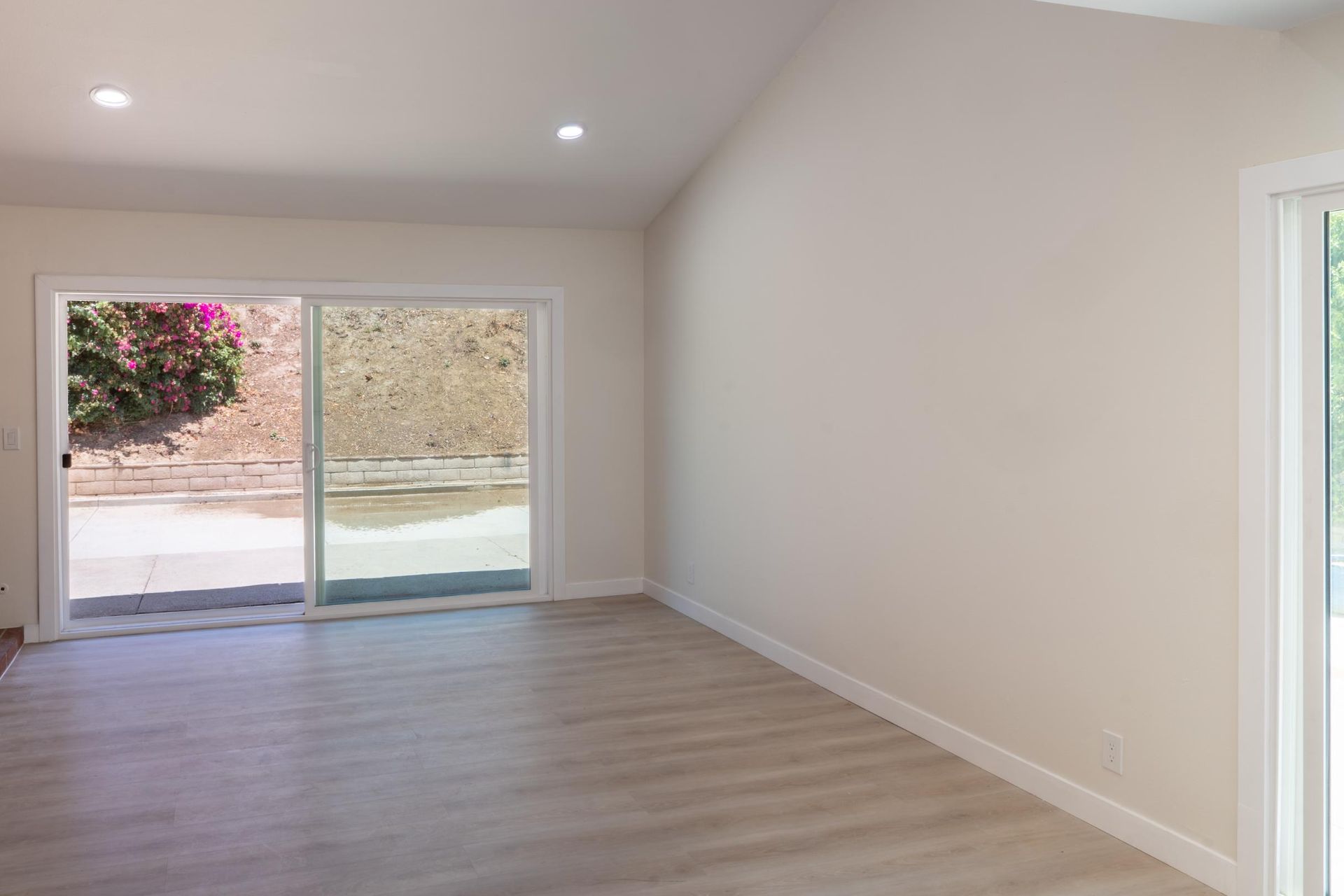 Empty bright room with light wood floors, recessed lights, and sliding glass doors to a patio.