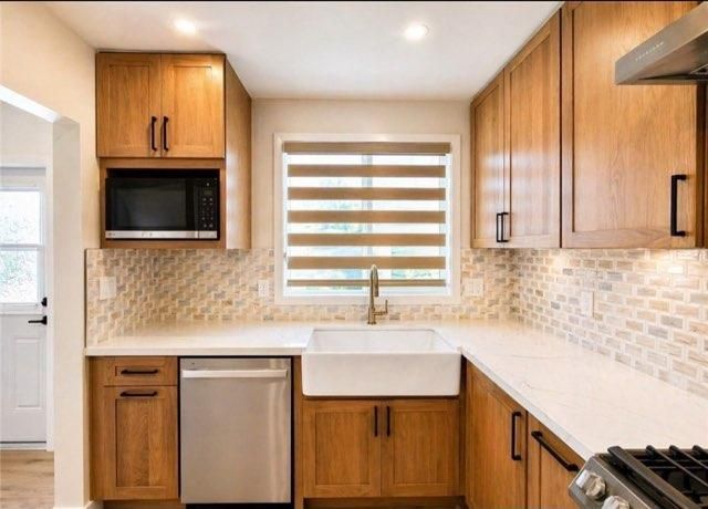 Modern kitchen with wood cabinets, white countertops, tiled backsplash, and a farmhouse sink under a window.