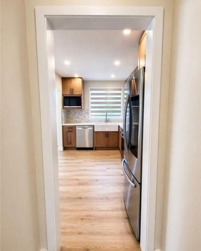 Narrow view into a bright kitchen with stainless steel appliances, wood cabinets, and light wood floors.
