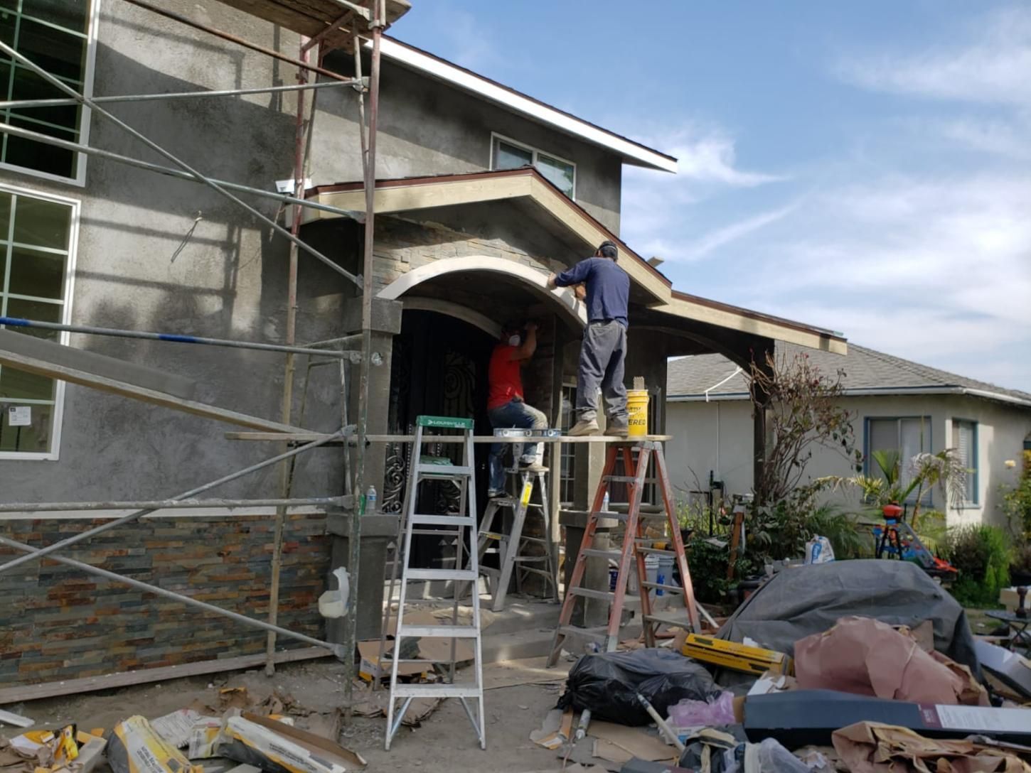 Workers on ladders repairing the front entrance of a house, with scaffolding and construction debris nearby