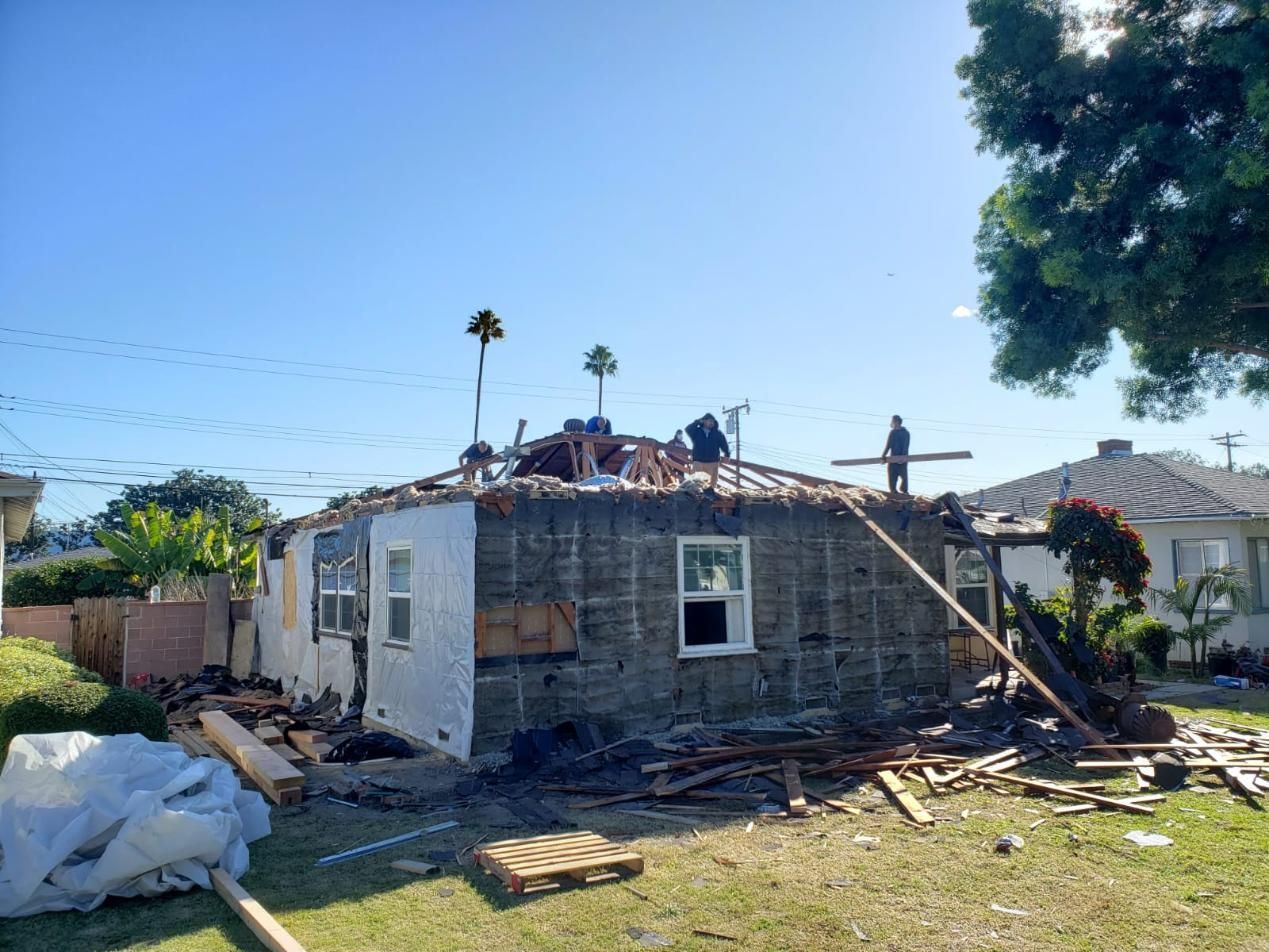 Damaged single-story house with collapsed roof and debris scattered in the yard under a clear blue sky