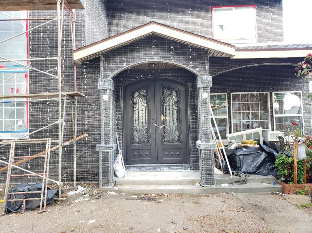 Front entrance of a house under construction with black double doors, scaffolding, and building materials nearby