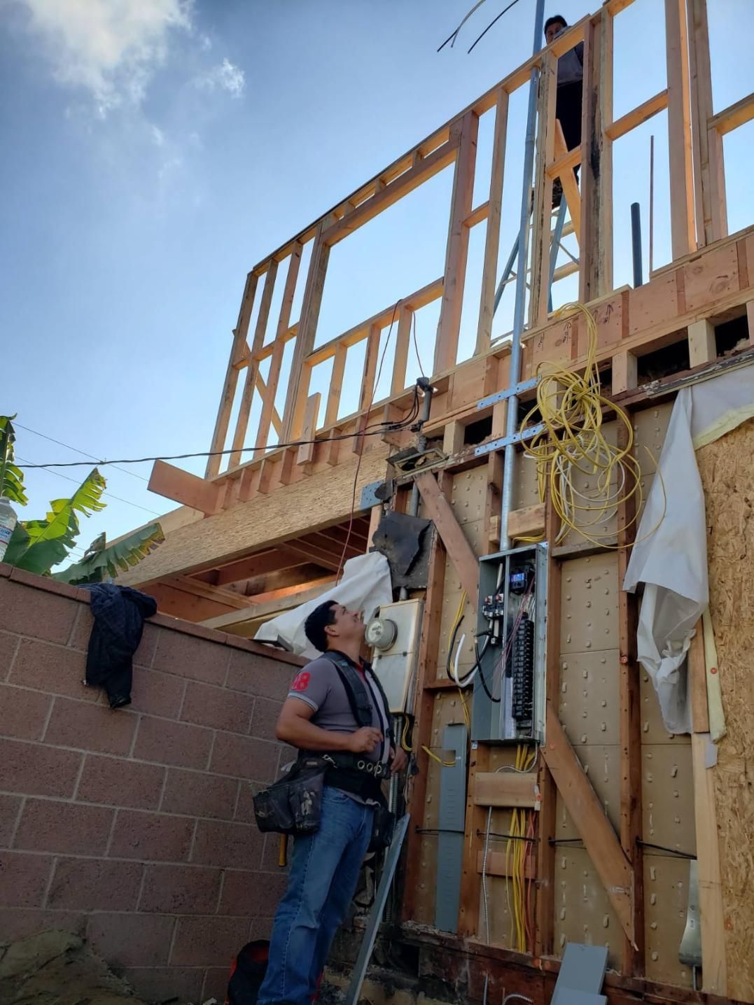 Two workers on a wooden house frame, one on a ladder and one below, with tools and materials visible.