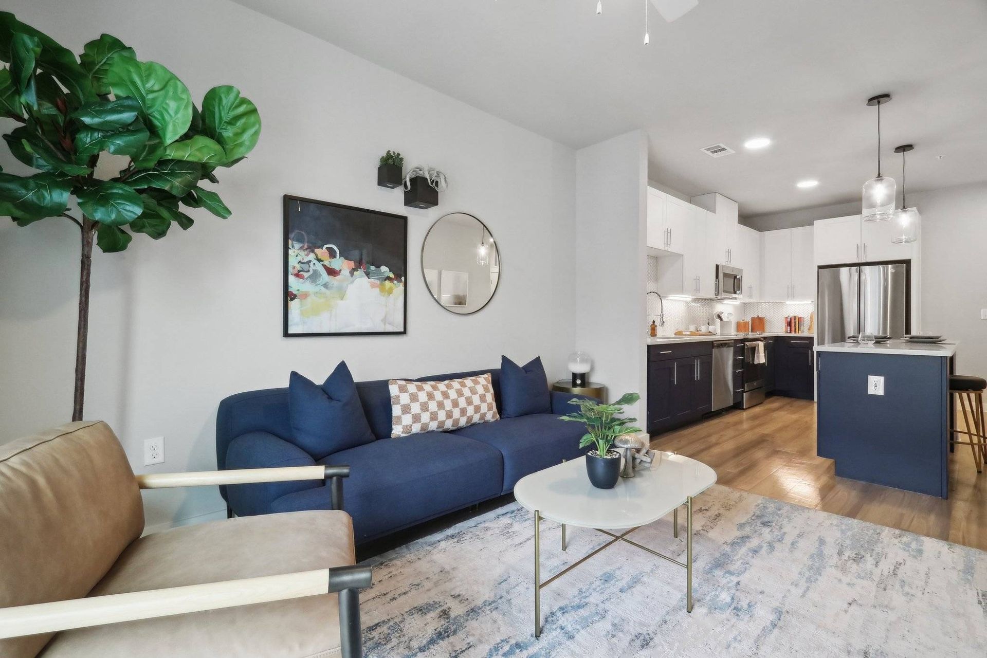 Open-concept living room with a blue sofa, plant, wall art, and a modern kitchen in the background.