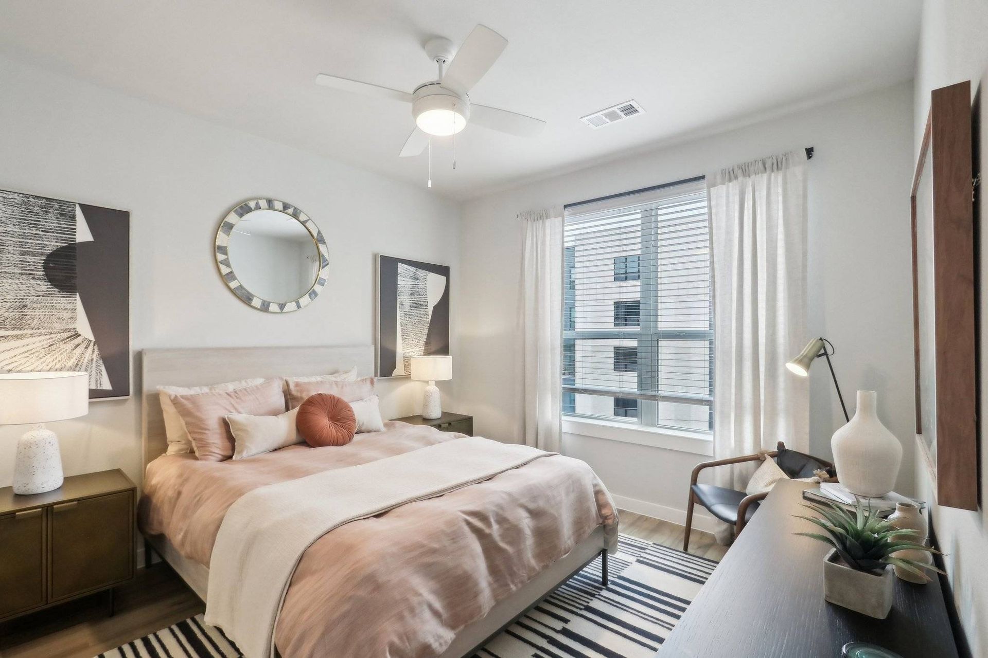 Bedroom in an apartment with a bed, round mirror, two nightstands, and a window with blinds.