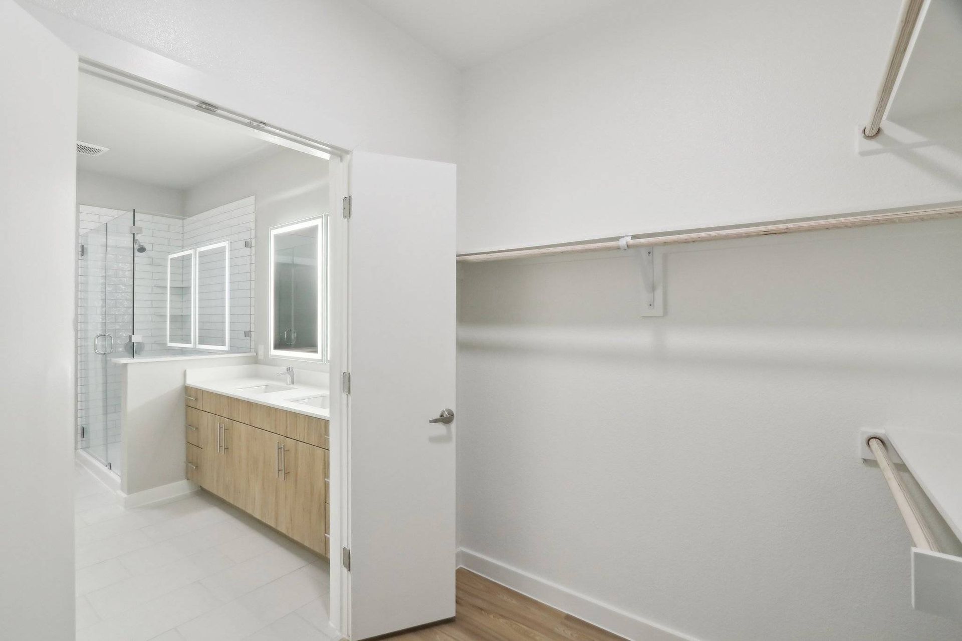 Modern apartment bathroom with double-sink vanity, glass-enclosed shower, and adjacent closet rod.