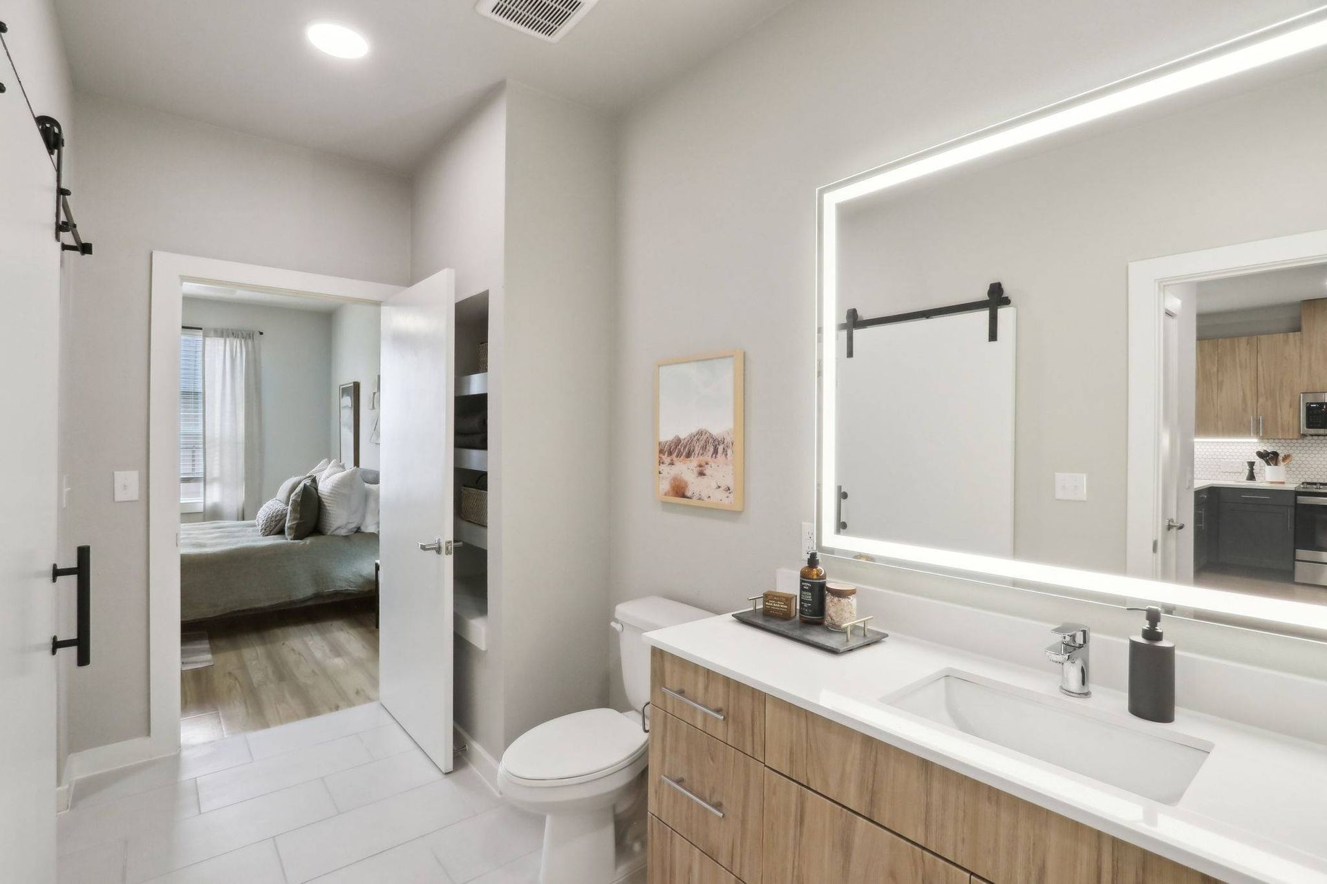 Bathroom with a wood-tone vanity, large backlit mirror, and open door to a bedroom closet.