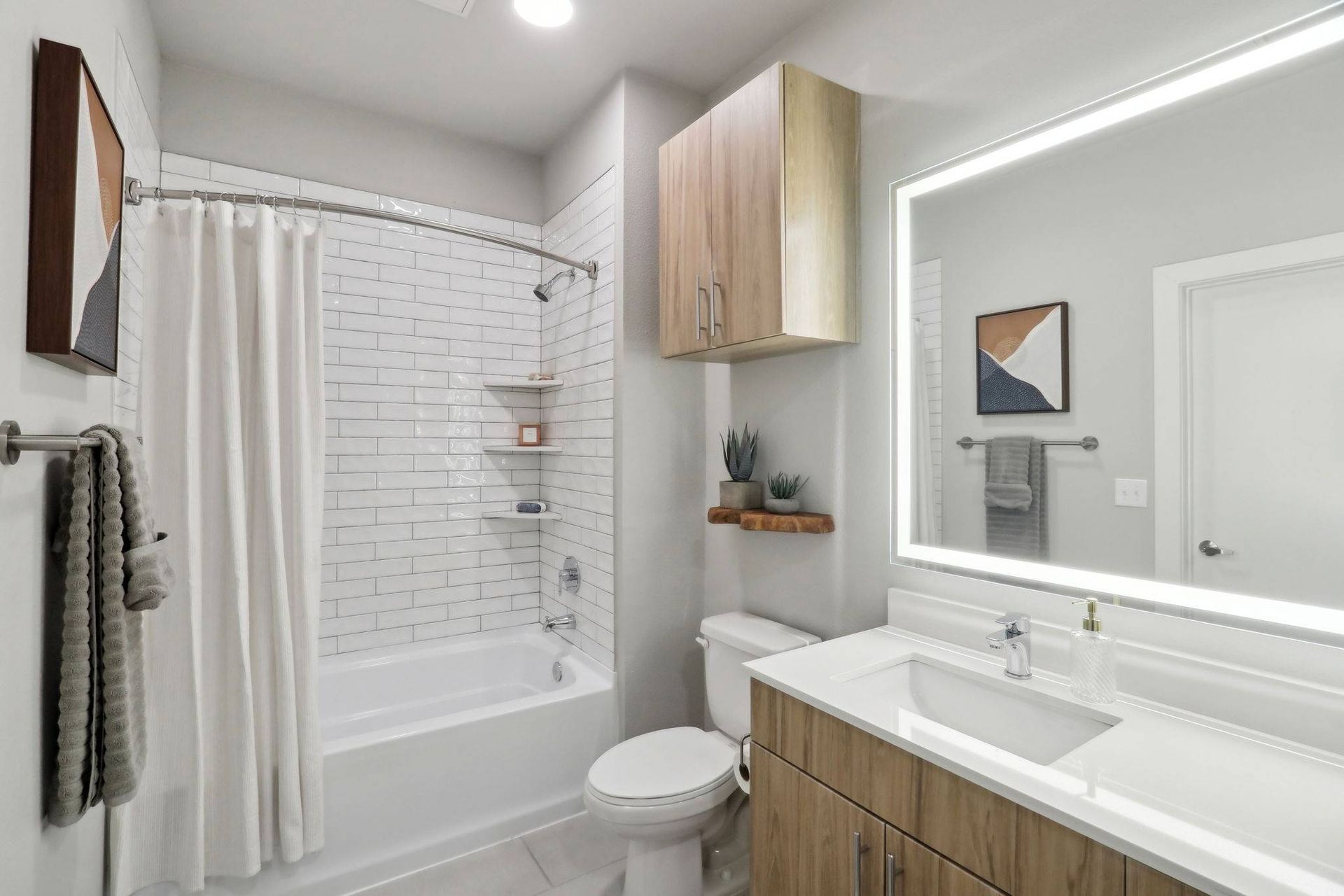 Bathroom in an apartment with a tub/shower, white subway tiles, a wood vanity, and a backlit mirror.