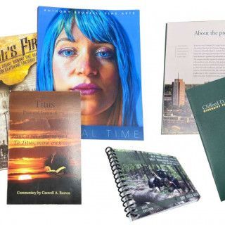 Assorted books and magazines fanned out on a white surface, including a blue-haired portrait cover and spiral notebook