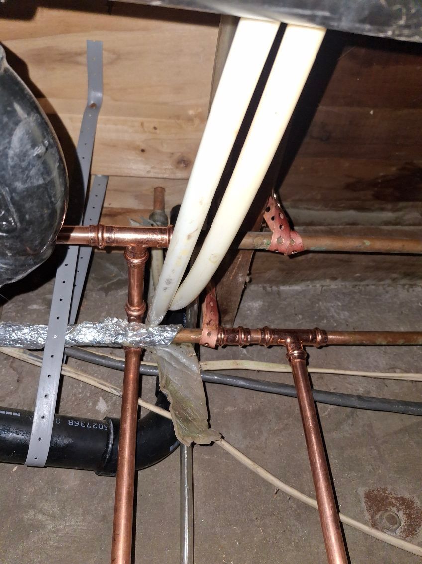 Exposed copper pipes and white PEX tubing under a wooden floor with insulation and a drain line
