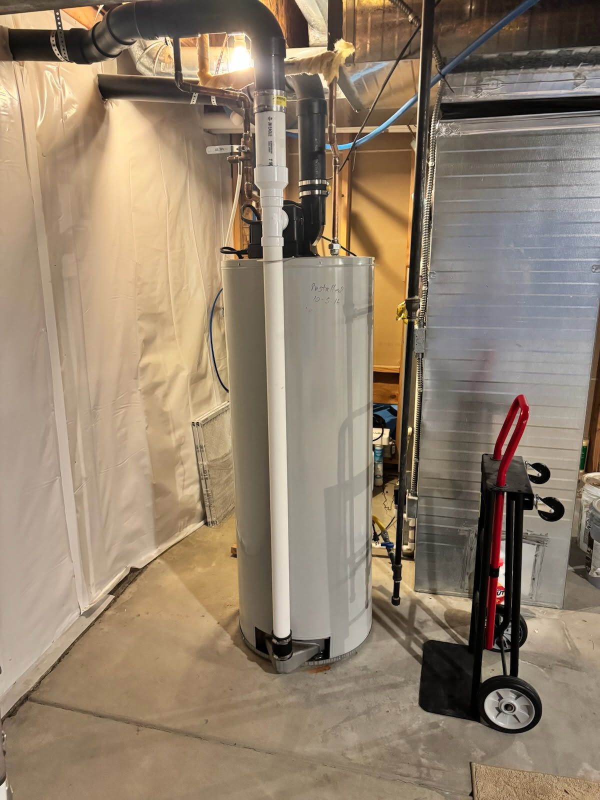 Water heater in a basement utility area with exposed pipes and a red hand truck nearby