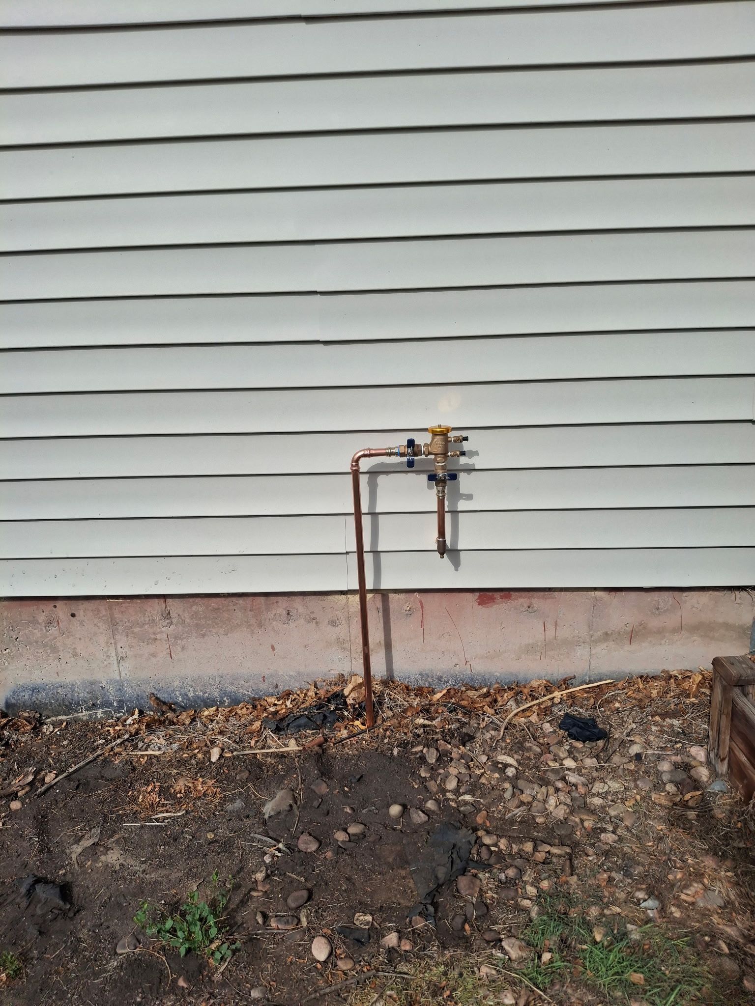 Outdoor water spigot and exposed pipes beside a house with white siding and bare ground below