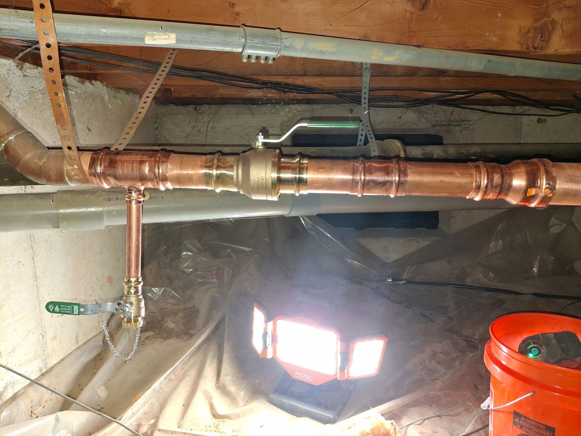 Copper plumbing pipes and valves under a basement ceiling with a red bucket and window light