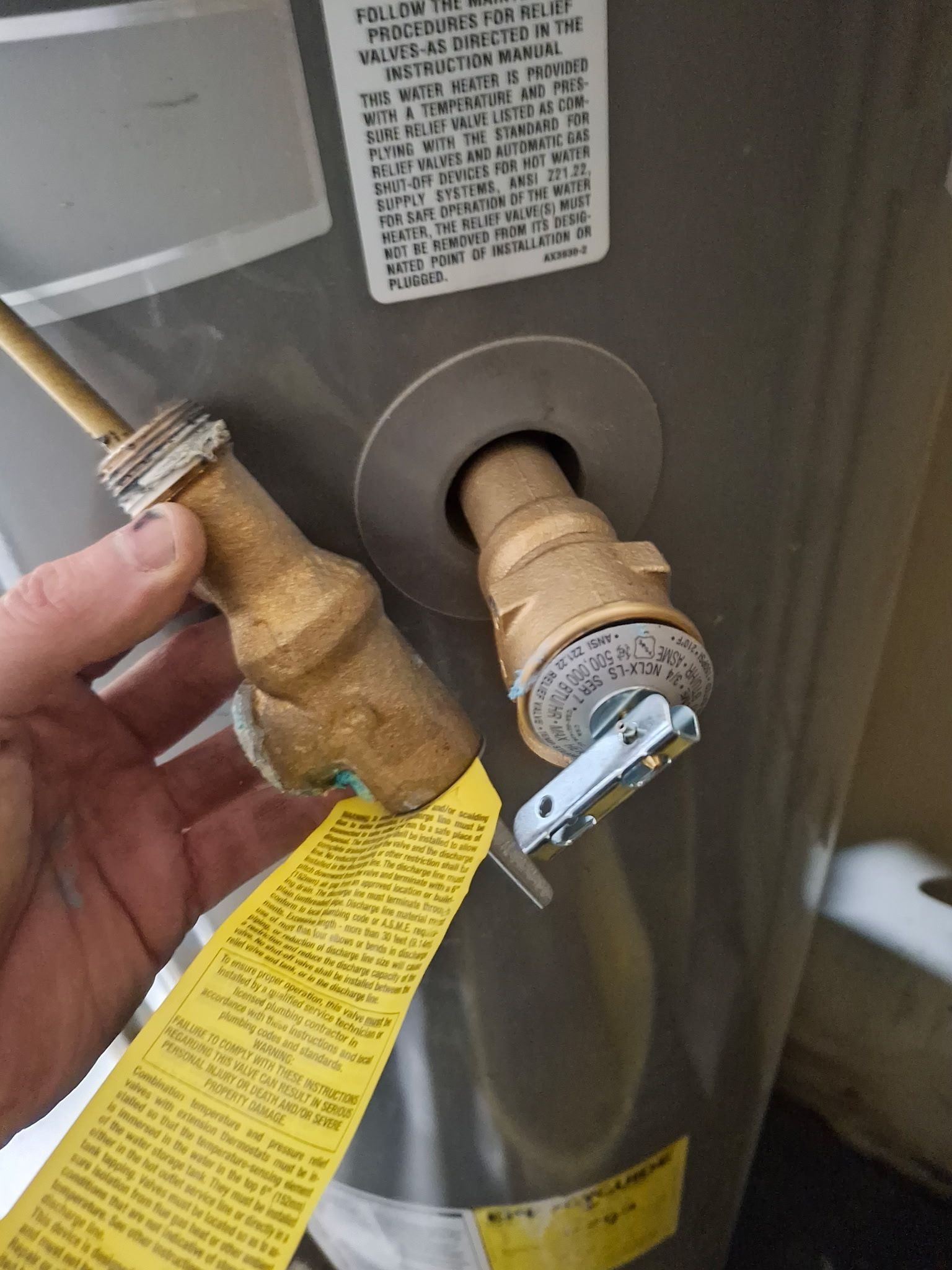 Hand holding a brass gas valve beside a water heater with a yellow warning tag.